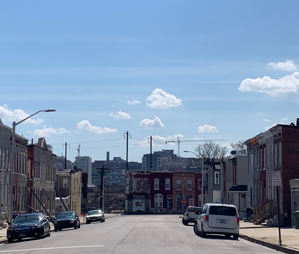 Picture of a street that dead ends into  deteriorating row homes with a blue sky above it and Johns Hopkins Hospital in the background.