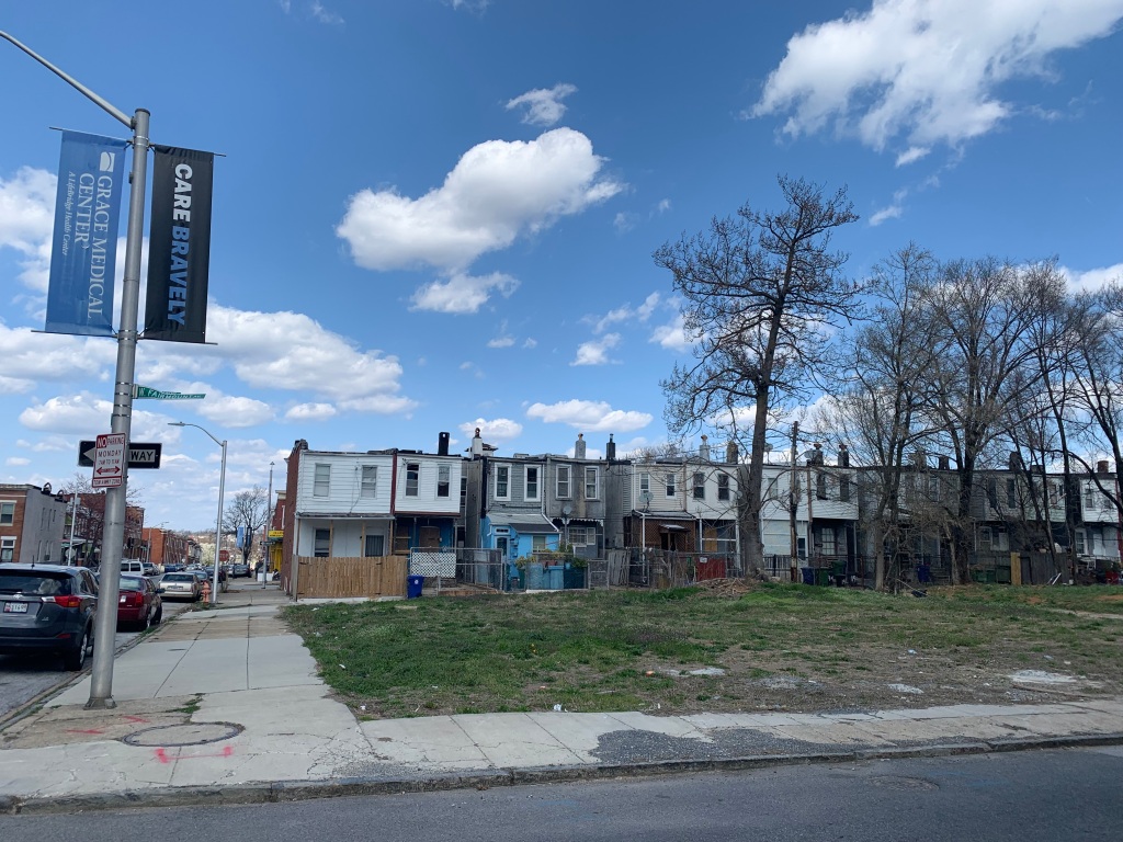 Picture of an empty lot in the foreground with row houses in the background. The sky is bright blue and dotted with clouds. On the left is a street light with flags reading "Grace Medical Center" on one side and "CARE BRAVELY" on the other side.